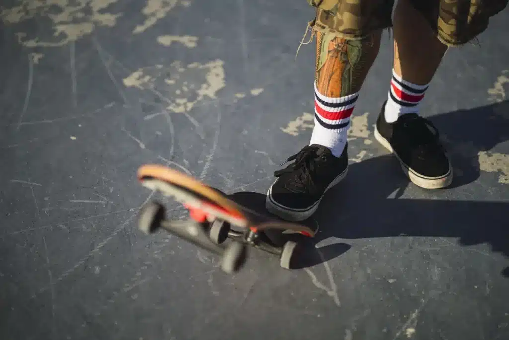 Pernas de skatista com bermuda camuflada, meia listrada e tênis pretos, posicionando um skate sobre superfície desgastada de pista