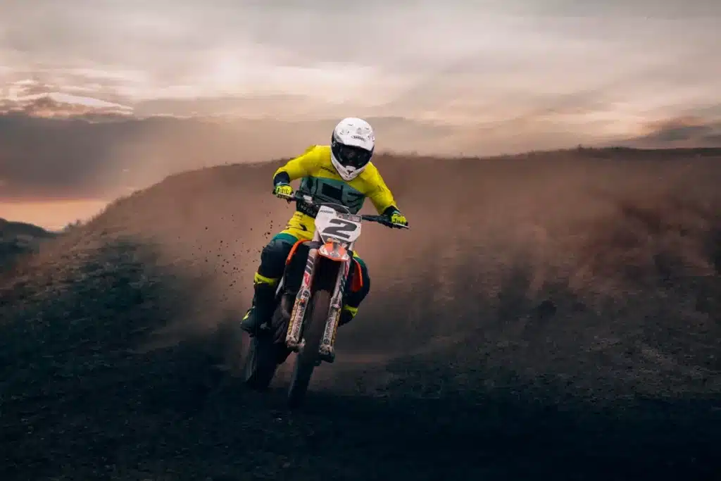 Criança em roupa de motocross e capacete pilotando uma moto número 2 em pista de terra, levantando poeira ao entardecer