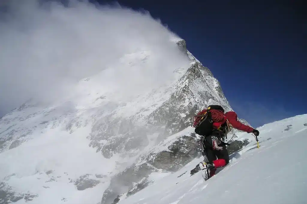 Um alpinista solitário, equipado com roupas de frio e mochila, escalando uma encosta nevada íngreme sob um céu azul intenso, com o pico da montanha coberto por nuvens no fundo, ilustrando a desafiadora prática do alpinismo.