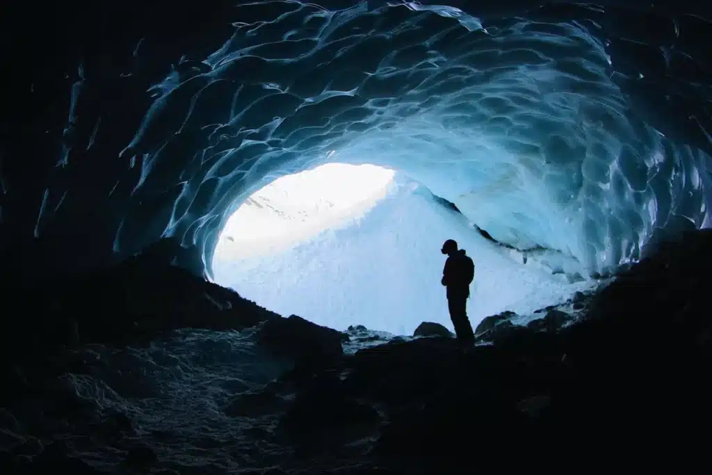 Silhueta de um escalador com capacete e piolets dentro de uma deslumbrante caverna de gelo azul, olhando para a saída brilhante, representando a beleza e o desafio do ice climbing.