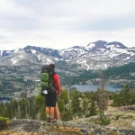 Homem com mochila de trekking apreciando paisagem de montanhas com neve e florestas ao fundo, em meio a uma trilha.