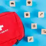 Kit de primeiros socorros vermelho acompanhado de pequenos blocos de madeira com ícones médicos sobre um fundo azul.