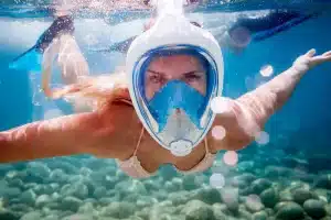 Mulher praticando mergulho com snorkel de rosto inteiro em água cristalina com fundo de pedras.