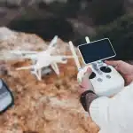 Pessoa operando um drone com controle remoto e tela integrada, em um ambiente externo rochoso.