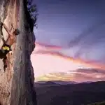Homem praticando escalada em uma parede rochosa íngreme ao ar livre, com o céu ao fundo em tons de roxo, rosa e laranja durante o pôr do sol.