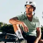 Homem com capacete de proteção branco, sentado ao lado de uma bicicleta BMX em um ambiente ao ar livre, com expressão pensativa. Ele veste uma camiseta verde clara e shorts escuros.