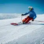 Esquiador descendo uma pista de neve com roupa azul e calça vermelha em um dia ensolarado com céu azul.