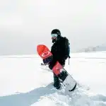 Pessoa praticando snowboard em uma paisagem coberta de neve, segurando a prancha vermelha com design moderno, equipada com capacete e óculos de proteção.