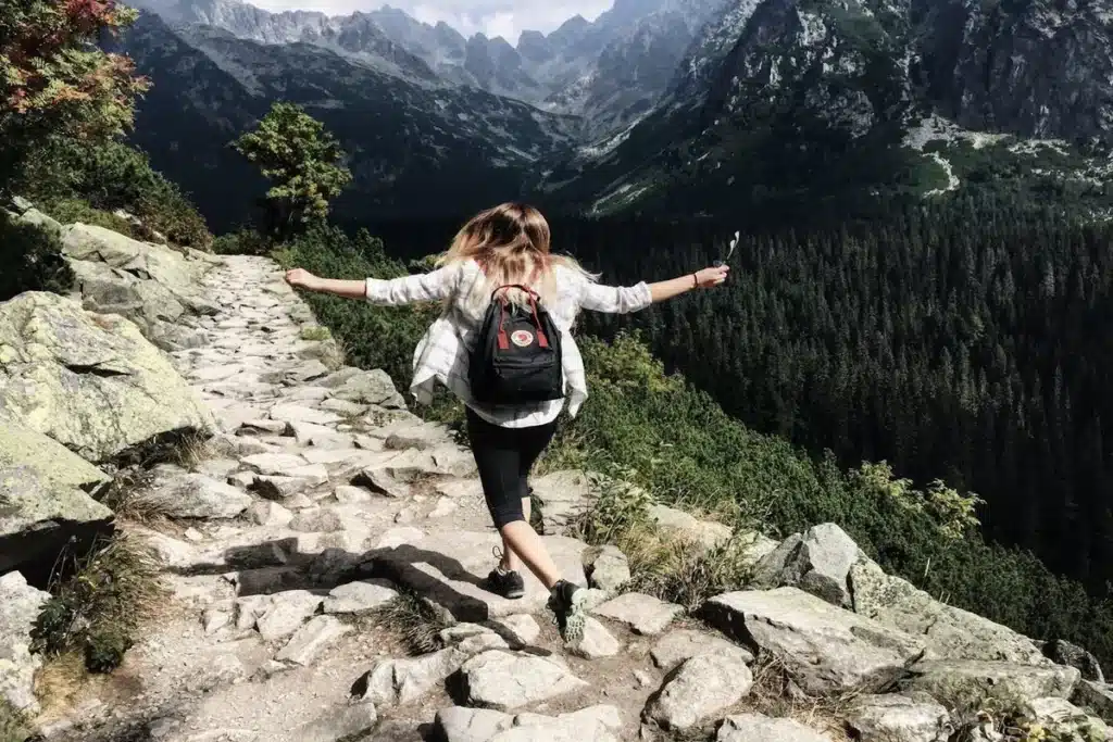 Mulher caminhando por uma trilha de pedras em uma montanha com os braços abertos, apreciando a paisagem de picos rochosos e floresta de pinheiros, simbolizando a liberdade e a aventura da trilha.
