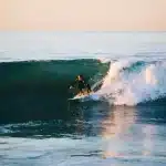 Surfista deslizando sobre uma onda no oceano, com a luz dourada do pôr do sol refletindo na água.