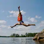 Mulher usando colete salva-vidas laranja salta de um rochedo para um lago, com os braços e pernas estendidos no ar.