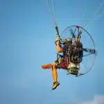 Piloto de paramotor voando no céu azul com equipamento motorizado e parapente colorido.