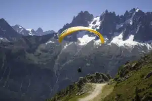 Pessoa praticando parapente com asa amarela sobre montanhas nevadas e paisagem rochosa, exibindo a emoção do voo livre.