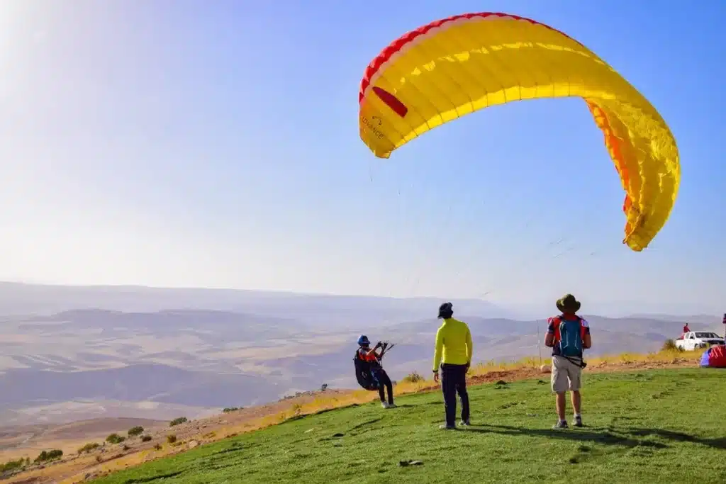 Pessoa decolando em parapente amarelo e vermelho de uma colina gramada, com paisagem montanhosa ao fundo, representando a liberdade do voo.