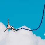 Homem saltando de bungee jump preso por corda elástica, com céu azul e nuvens ao fundo