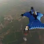 Praticante de wingsuit voando a grande altitude sobre uma paisagem rural, usando traje azul aerodinâmico para planar.