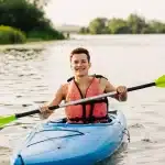Rapaz sorridente rema em um caiaque azul sobre as águas calmas de um lago, vestindo colete salva-vidas e cercado por vegetação natural.