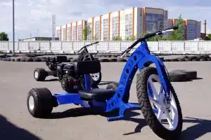 "Drift trike azul motorizado estacionado em pista de asfalto, com pneus de proteção e edifícios ao fundo"