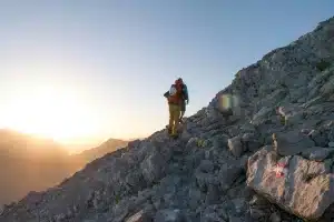 Dois montanhistas subindo trilha rochosa ao nascer do sol, com mochilas e equipamentos