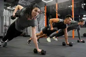 Homens e mulher em academia fazem exercício funcional em posição de prancha com halteres, demonstrando força, foco e sincronia.