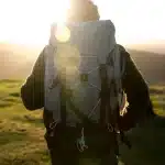 Pessoa com mochila de trilha observa o nascer do sol sobre as montanhas, em um cenário de natureza ao ar livre.