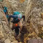 Grupo de montanhistas escalando passagem estreita e rochosa com capacetes e mochilas.