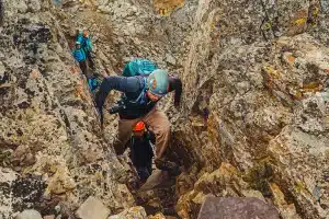 Grupo de montanhistas escalando passagem estreita e rochosa com capacetes e mochilas.