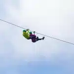 Duas pessoas praticando tirolesa em dupla, com roupas coloridas e capacetes, suspensas em cabo de aço no céu azul