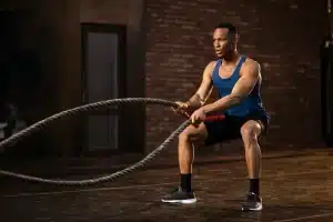 Homem com camiseta azul e shorts pretos executa exercício de corda naval em ambiente de treino funcional, demonstrando força e foco.