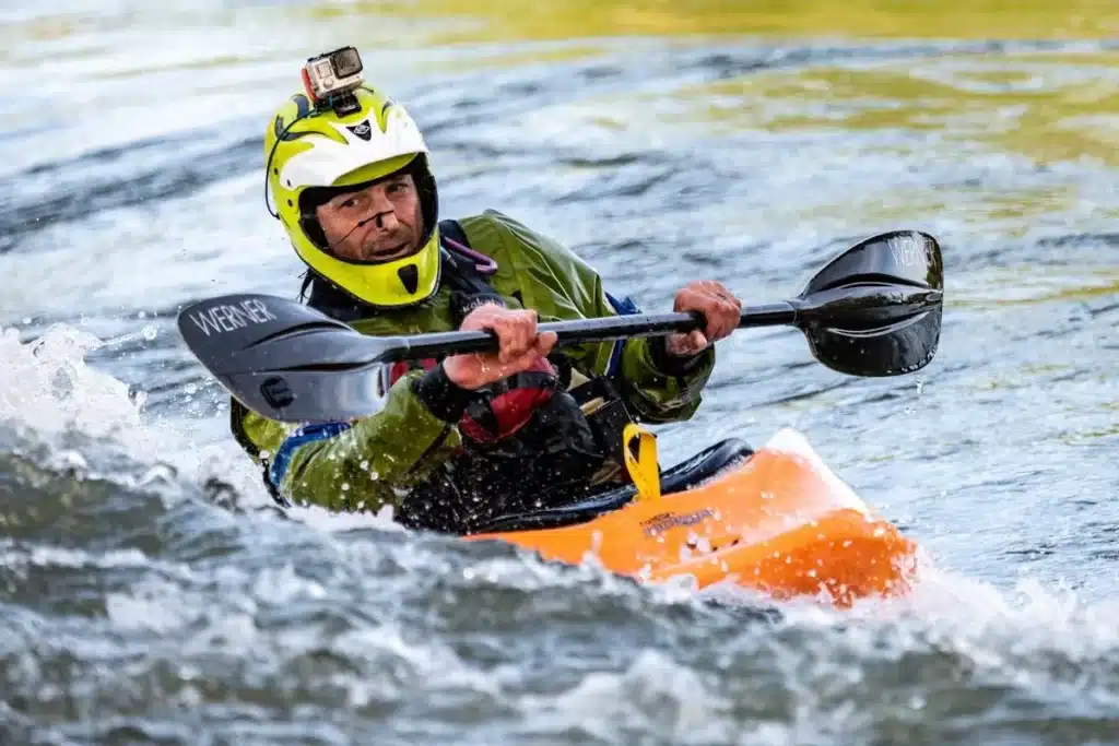 Um caiaquista com um capacete amarelo e verde, equipado com uma câmera "GoPro" montada no topo, rema vigorosamente em águas turbulentas, capturando a emoção do esporte.