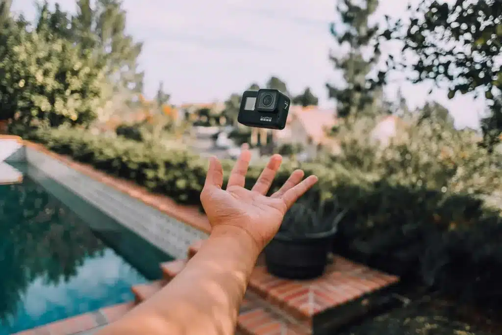 Mão de uma pessoa jogando uma câmera GoPro preta no ar próximo a uma piscina, com árvores e casas ao fundo em um ambiente externo iluminado pelo sol.