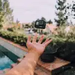Mão de uma pessoa jogando uma câmera GoPro preta no ar próximo a uma piscina, com árvores e casas ao fundo em um ambiente externo iluminado pelo sol.