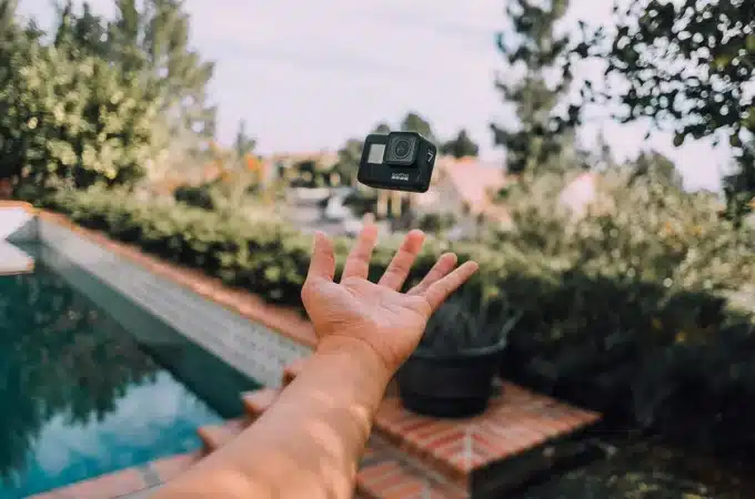 Mão de uma pessoa jogando uma câmera GoPro preta no ar próximo a uma piscina, com árvores e casas ao fundo em um ambiente externo iluminado pelo sol.