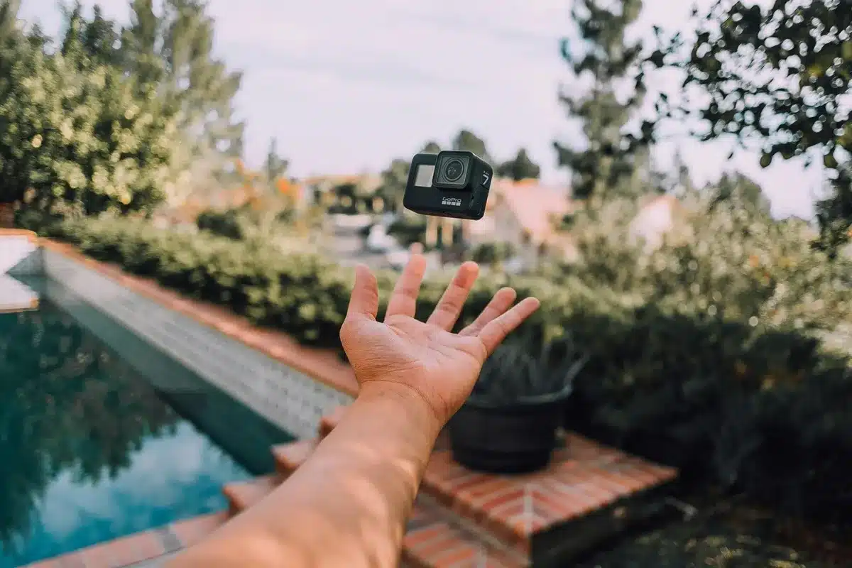 Mão de uma pessoa jogando uma câmera GoPro preta no ar próximo a uma piscina, com árvores e casas ao fundo em um ambiente externo iluminado pelo sol.
