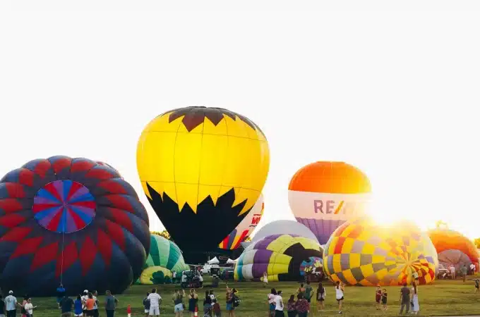 Imagem de balões de ar quente coloridos sendo preparados para voar, com espectadores ao fundo em um evento de balonismo.