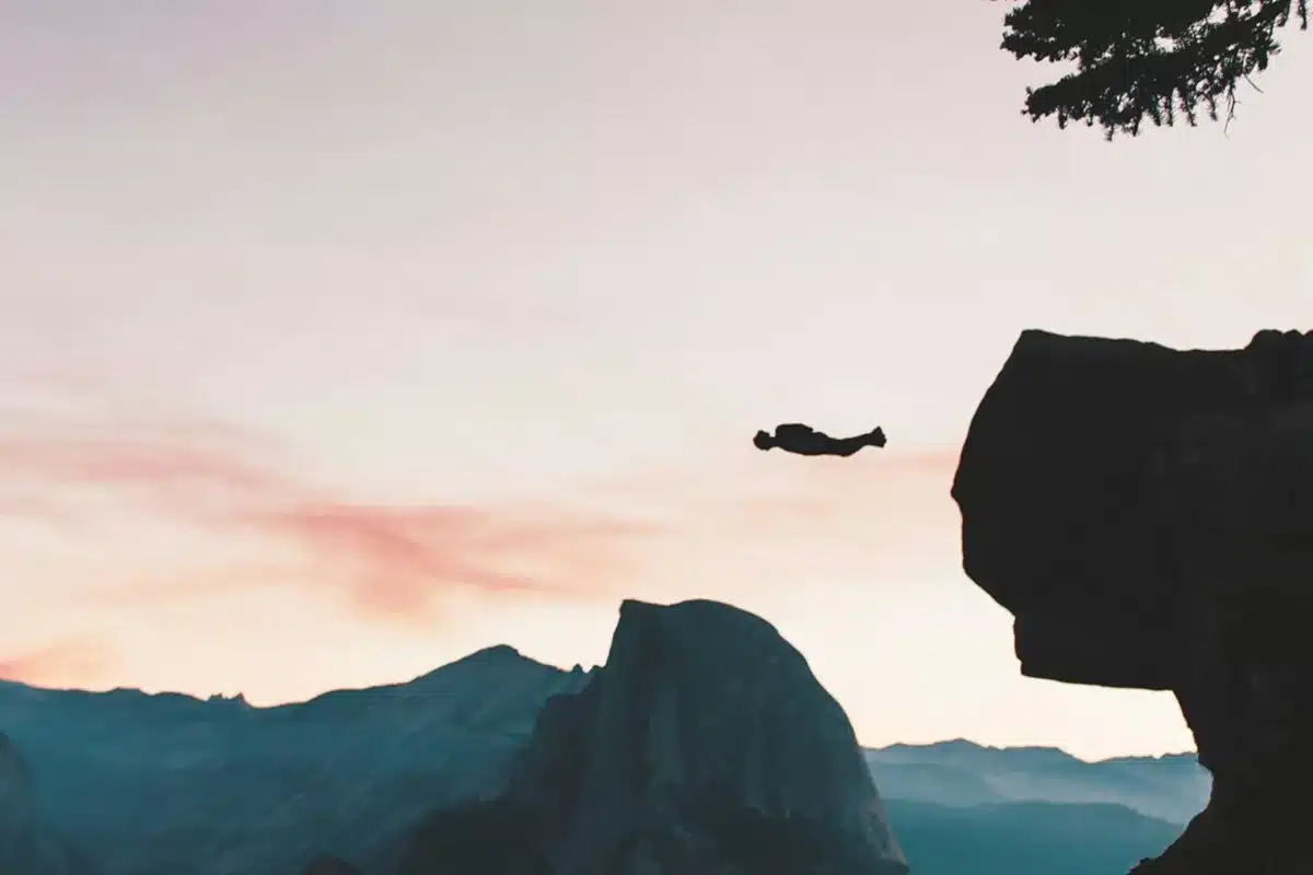 Pessoa realizando salto de base jump de um penhasco com o corpo na posição horizontal, ao entardecer, com montanhas ao fundo e céu em tons de rosa e laranja.