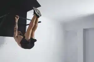Homem tatuado, sem camisa, realizando exercício de elevação de pernas em barra fixa, treinando calistenia em ambiente interno de paredes brancas.