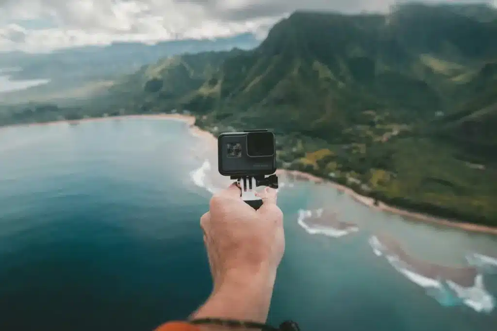 Mão de uma pessoa segurando uma câmera GoPro com fundo de vista aérea de um litoral com montanhas verdes, praia e mar azul, sob um céu parcialmente nublado.