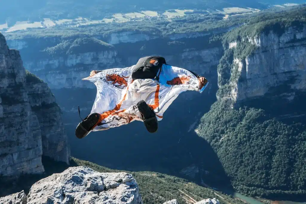 Pessoa usando traje wingsuit com estampa de tigres realizando salto de base jump de um penhasco alto, com cânion profundo e florestas ao fundo.