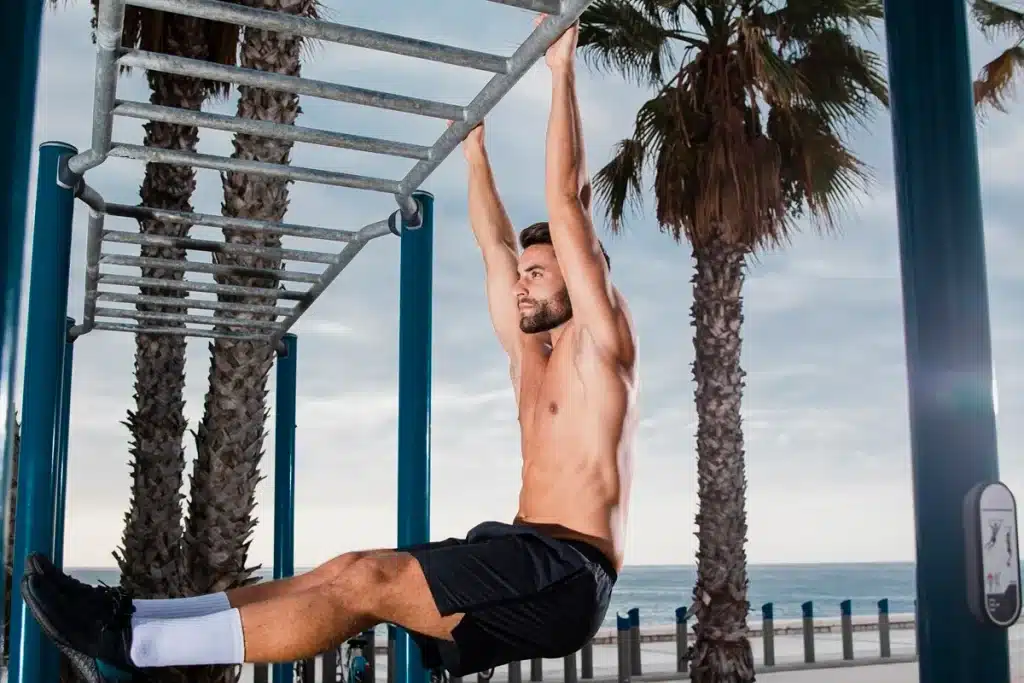 Homem sem camisa fazendo exercício de suspensão em barra fixa em parque de calistenia ao ar livre, com palmeiras e o mar ao fundo.