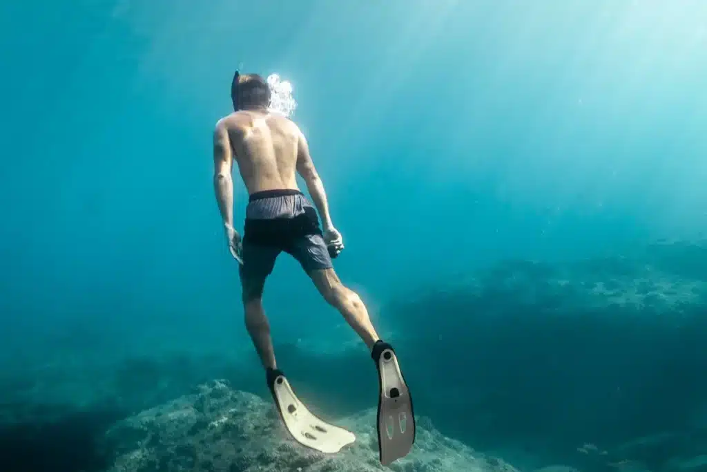 Imagem de um homem praticando mergulho por apneia, submergindo em águas claras com pés e mãos relaxados, enquanto bolhas sobem à superfície.