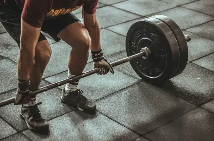 Imagem de um homem levantando um haltere pesado em uma academia, usando luvas de treinamento e com tênis de treino. O foco está na técnica de levantamento de peso, e o fundo é de piso de borracha.
