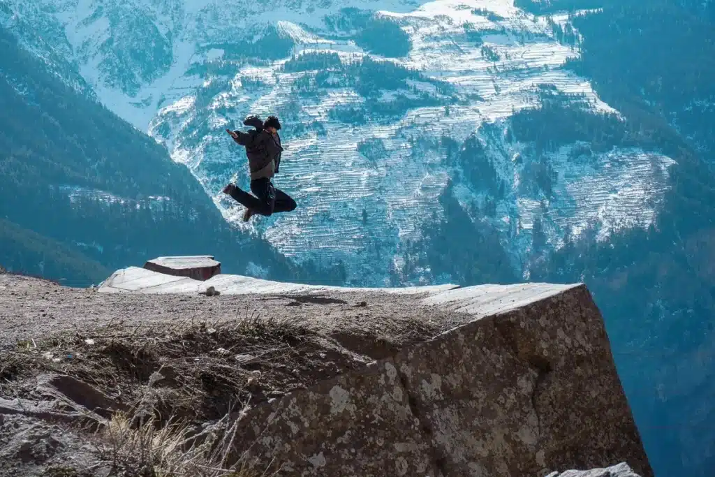 Pessoa saltando no topo de um penhasco com vista para montanhas cobertas de neve, simbolizando liberdade e aventura na natureza.