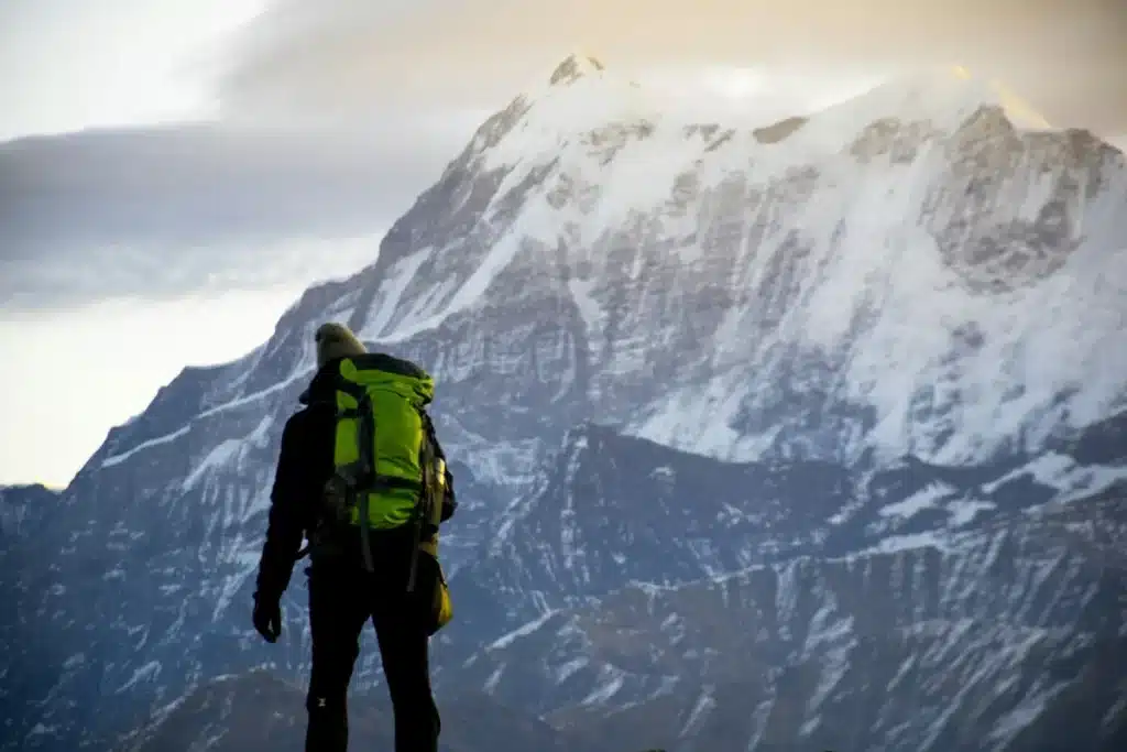 Alpinista com mochila verde fluorescente observando uma montanha coberta de neve ao entardecer.
