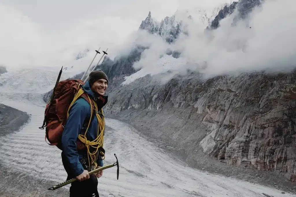 Alpinista sorridente com mochila e equipamentos de escalada diante de uma paisagem montanhosa nevada.