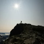 Alpinista solitário no topo de uma montanha rochosa sob o sol intenso em um céu claro.