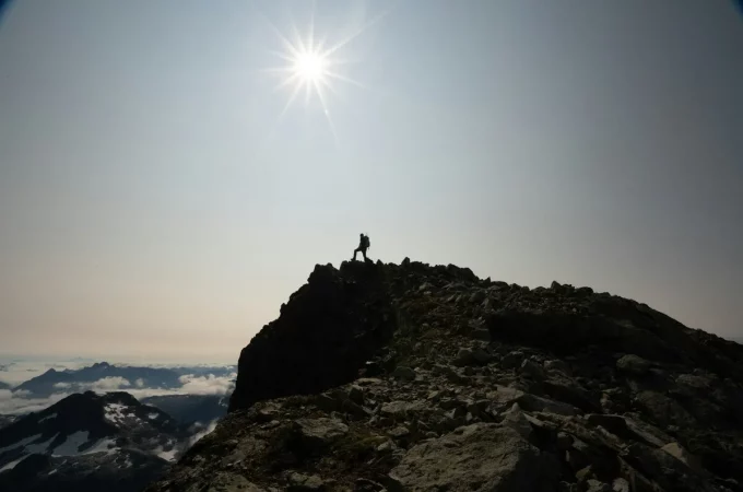 Alpinista solitário no topo de uma montanha rochosa sob o sol intenso em um céu claro.
