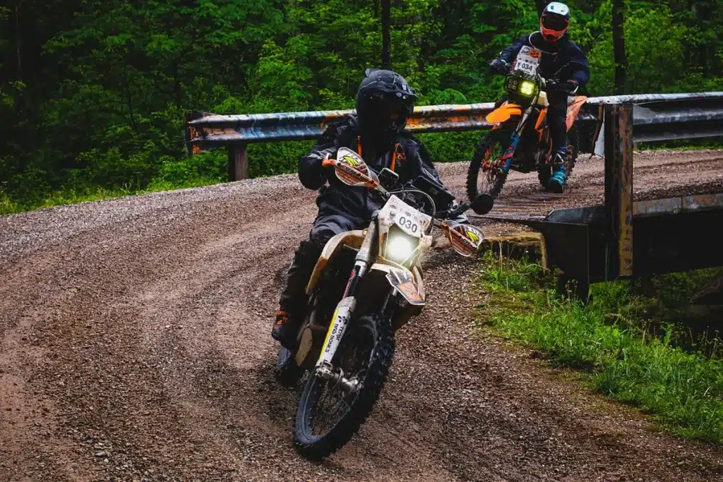 Dois motociclistas em uma competição de rally de moto em uma estrada de terra, passando por uma curva perto de uma ponte de metal.