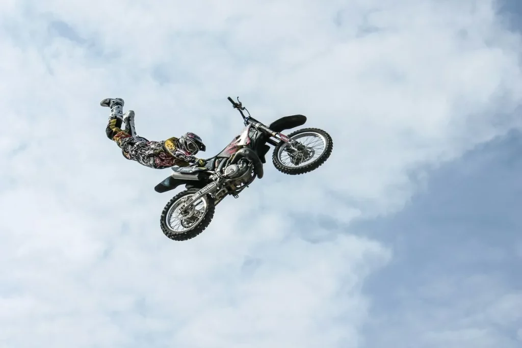 Piloto de motocross realizando manobra aérea de estilo livre, completamente no ar junto à moto contra o céu azul com nuvens