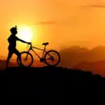 Ciclista de mountain bike em silhueta contra um pôr do sol alaranjado, com montanhas ao fundo, simbolizando a aventura do MTB.